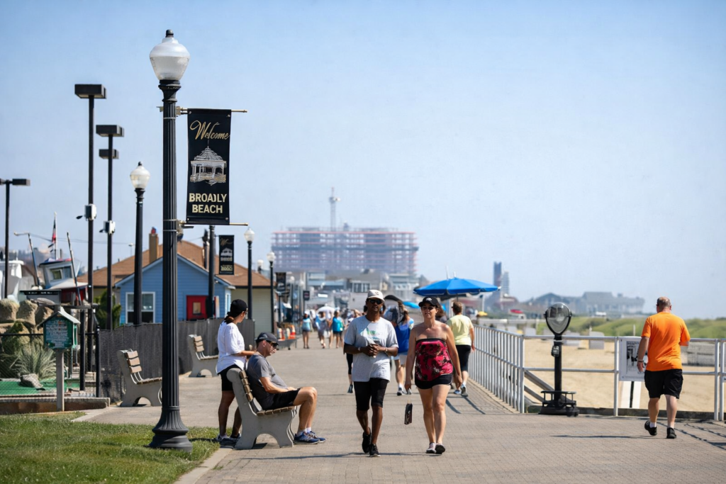 People walking along the Bradley Beach boardwalk on a sunny day, with a Welcome to Bradley Beach sign, benches, street lamps, and the sandy shoreline visible nearby