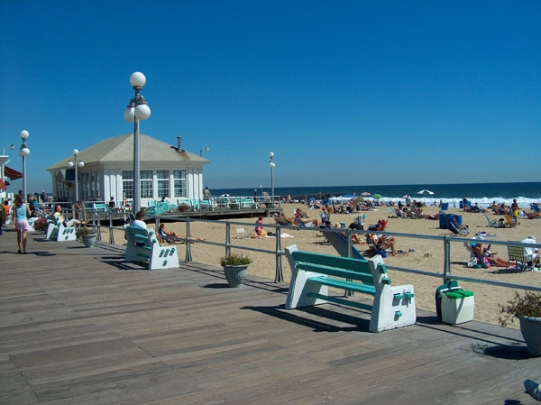 Sunny boardwalk promenade in Avon-by-the-Sea, NJ, with a classic white pavilion, green benches, and a crowded beach stretching along the Atlantic Ocean on a clear summer day