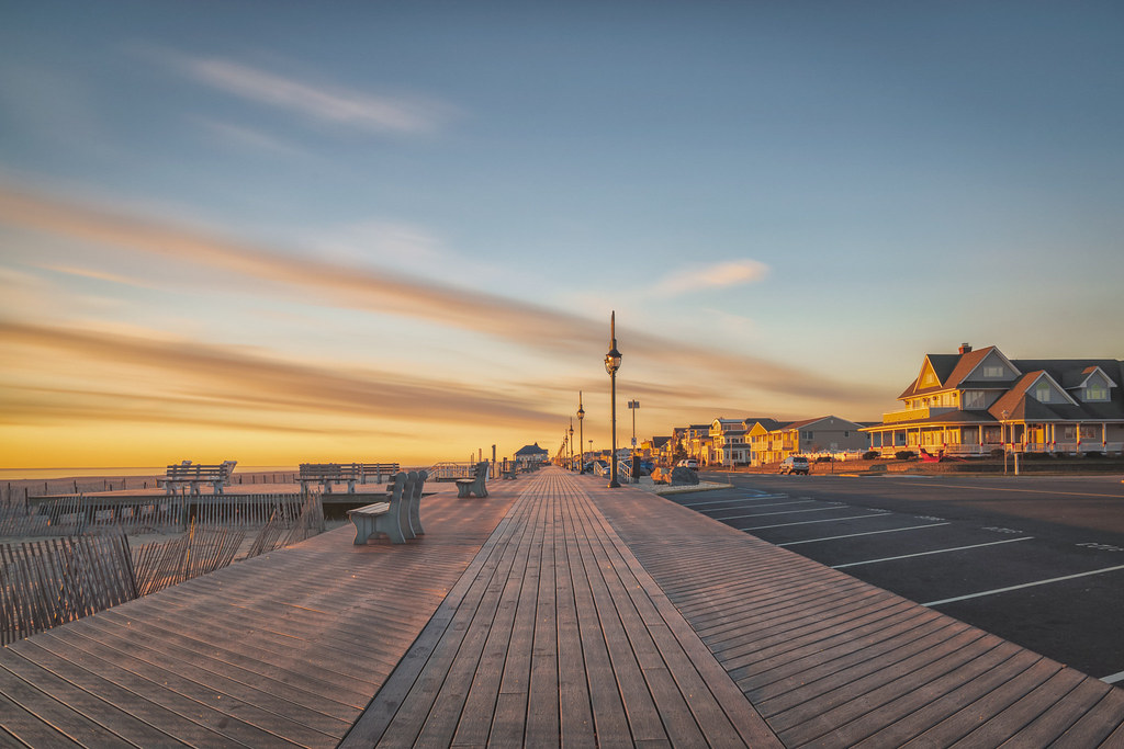 Empty Belmar boardwalk at sunset, with warm golden and pink skies reflecting over the Atlantic Ocean, benches lining the wide wooden promenade, and coastal homes glowing in the evening light