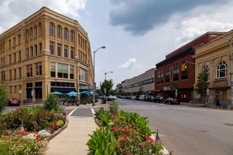Vibrant downtown Asbury Park, NJ, with historic mixed-use brick buildings, outdoor café seating, colorful flower beds, and active street life under a partly cloudy sky