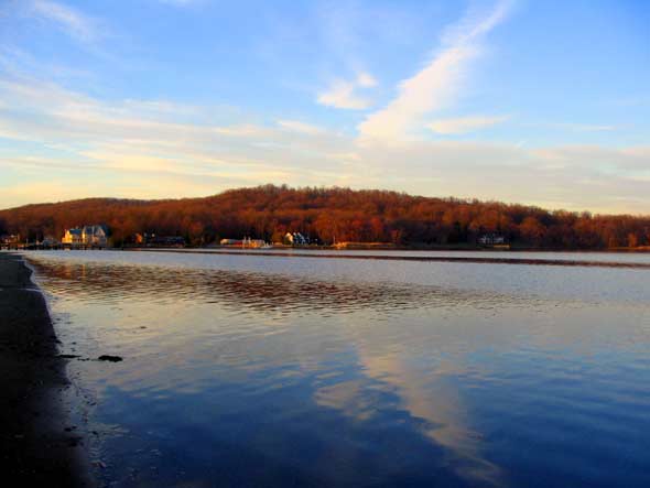 Peaceful winter view of the Navesink River in Rumson, NJ, with calm reflective waters, a sandy shoreline, and elegant estate homes nestled among auburn tree-covered hills across the river