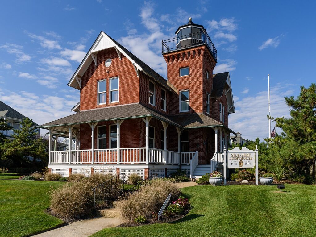The historic Sea Girt Lighthouse, a charming red brick Victorian structure with a wraparound porch and light tower, surrounded by manicured lawns in Sea Girt, NJ