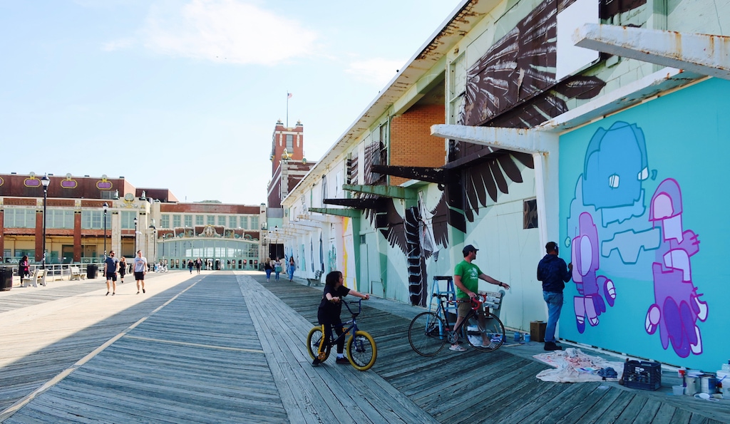 People walking and cycling along the Asbury Park boardwalk beside a large colorful street mural, with historic buildings visible in the background