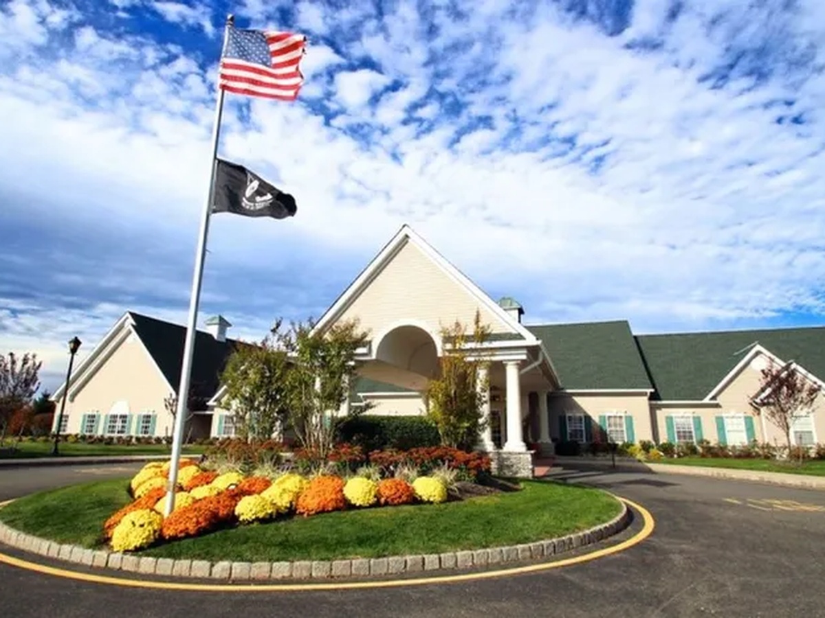 Welcoming clubhouse entrance of a 55+ active adult community in Toms River, NJ, with an American flag, colorful flower beds, and a circular driveway under a blue sky