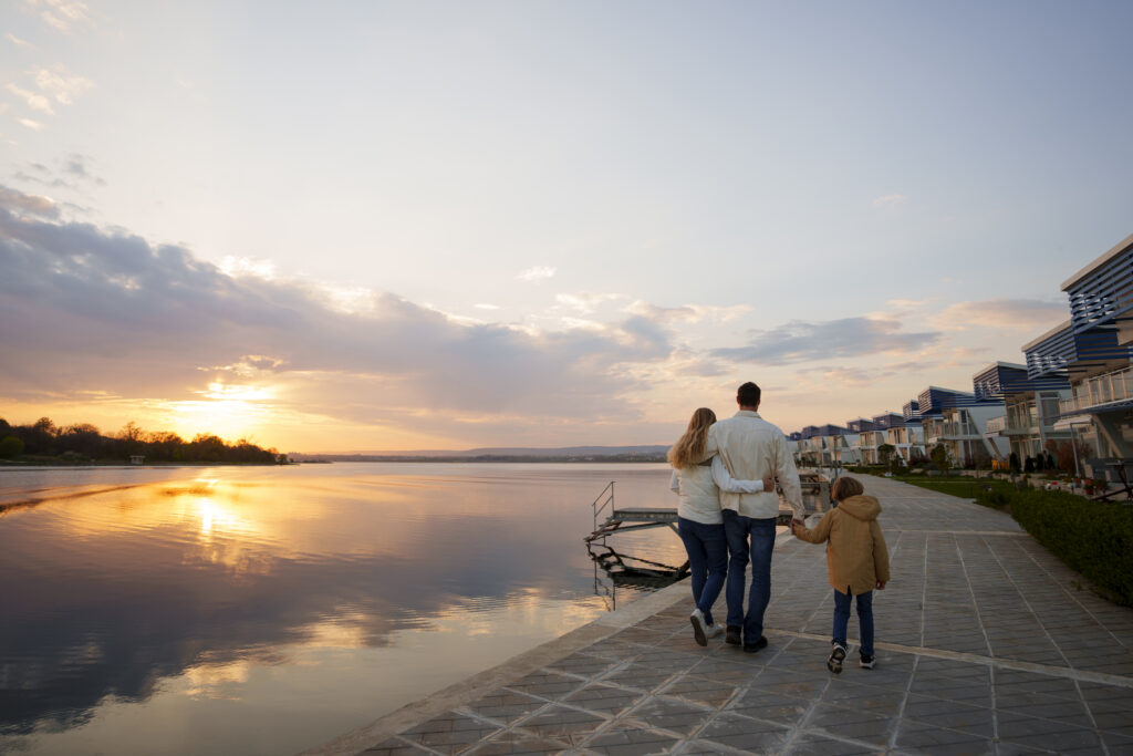 Young family strolling along a waterfront promenade at sunset in a Monmouth County, NJ riverfront community, with golden light reflecting on the calm river and residential homes lining the path