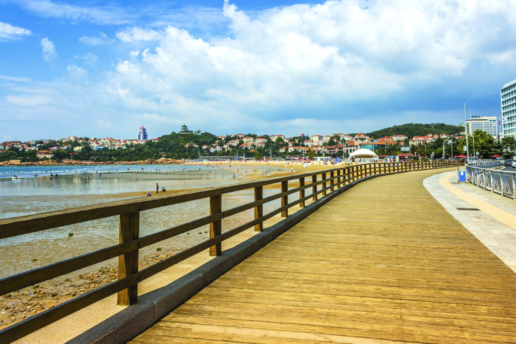 Wooden boardwalk promenade stretching along a sandy beach in a Monmouth County, NJ shore town, with calm ocean waters, a sandy shoreline, and a tree-covered coastal town in the background