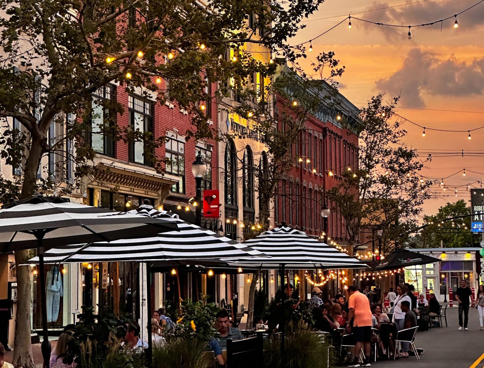 Lively outdoor dining scene in downtown Red Bank, NJ, with string lights, striped umbrellas, and brick buildings glowing under a warm sunset sky