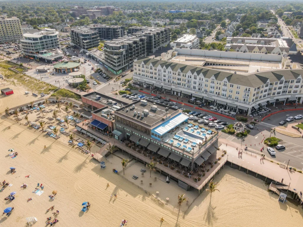 Aerial view of Long Branch's modern beachfront development with upscale hotels, mixed-use buildings, outdoor dining, and beachgoers enjoying the Atlantic Ocean on a summer day