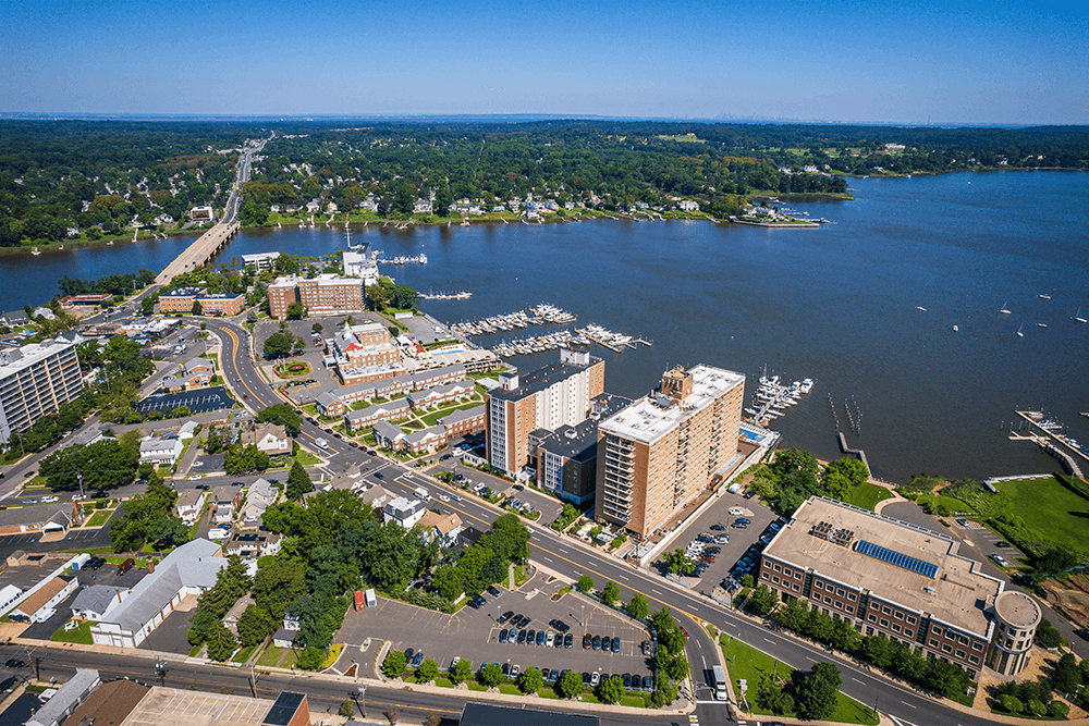 Aerial summer view of Red Bank, NJ, with a dense downtown core, mid-rise buildings, marinas with docked boats, and the wide Navesink River curving through lush green surroundings