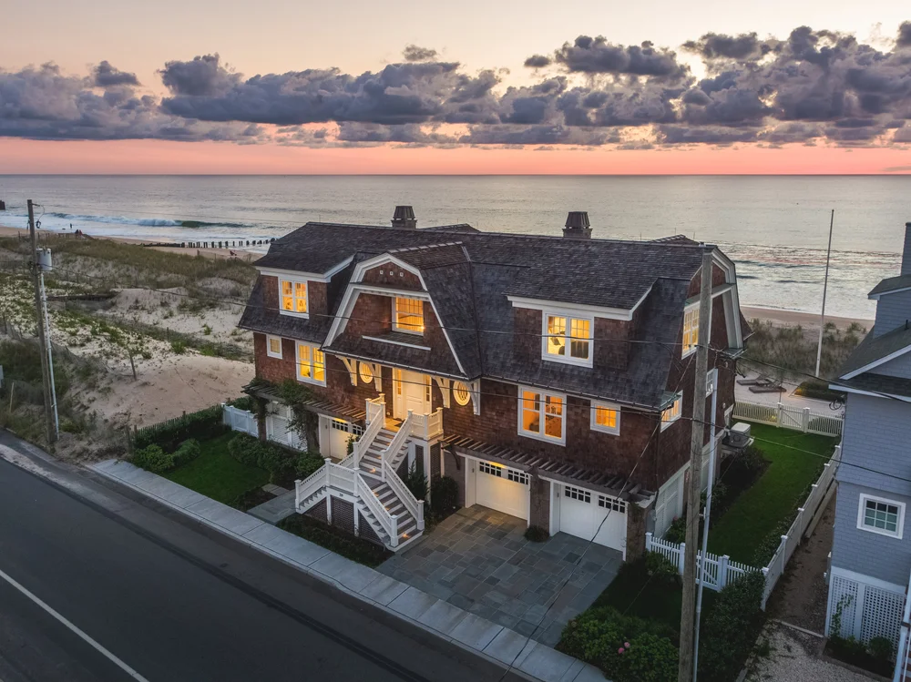Classic shingled oceanfront home in Bay Head, NJ, with glowing interior lights, a wide sandy beach, and a dramatic pink and purple dusk sky over the Atlantic Ocean