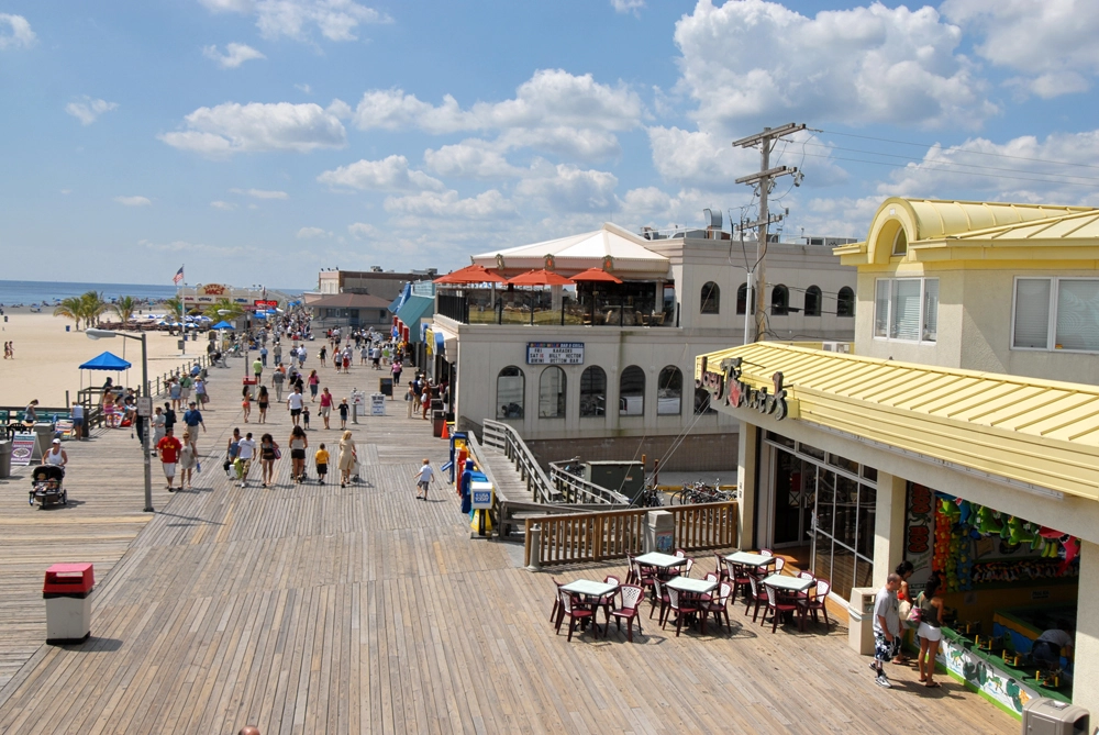 Busy boardwalk promenade in Point Pleasant Beach, NJ, with waterfront shops, outdoor dining, and families strolling along the oceanfront on a sunny summer day