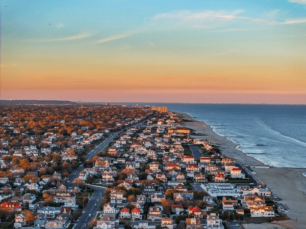 Aerial twilight view of Neptune Township, NJ, with rows of oceanfront homes glowing in warm sunset colors along the Atlantic shoreline and calm bay waters stretching inland