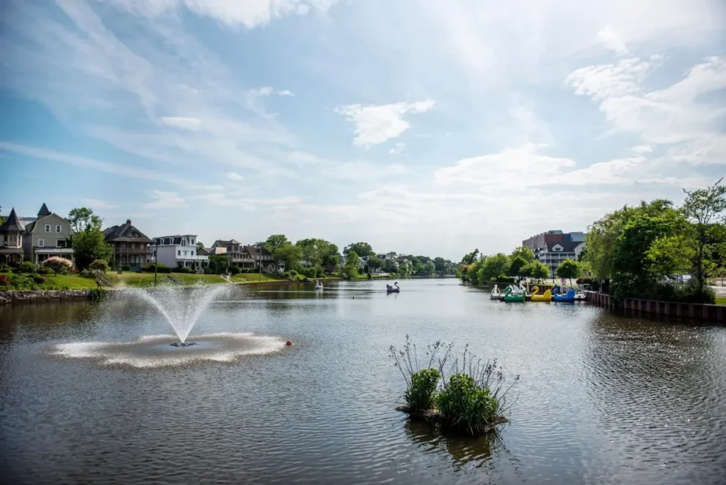 Peaceful lakeside scene in Interlaken, NJ, with a decorative fountain, colorful paddleboats, tree-lined shores, and charming homes reflecting on the calm waters of Deal Lake