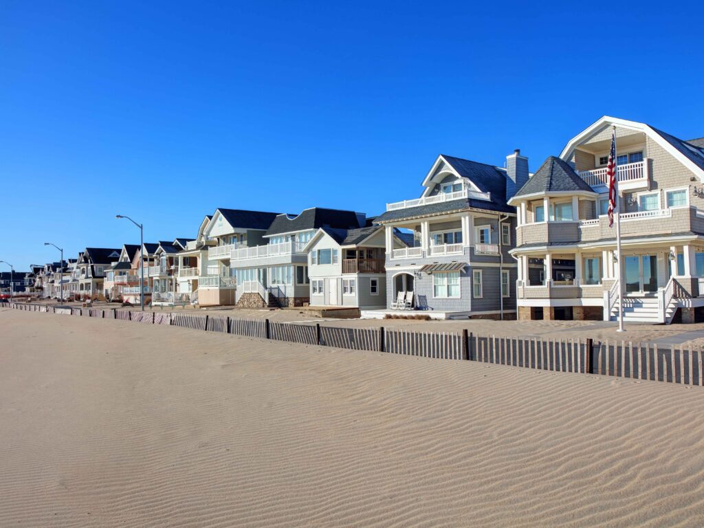 ow of charming beachfront homes with wraparound porches and an American flag along the sandy shoreline in Manasquan, NJ, under a bright blue sky